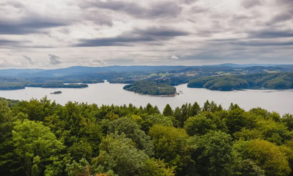 Widok na jezioro solińskie otoczone zielonymi wzgórzami i lasami pod pochmurnym niebem.