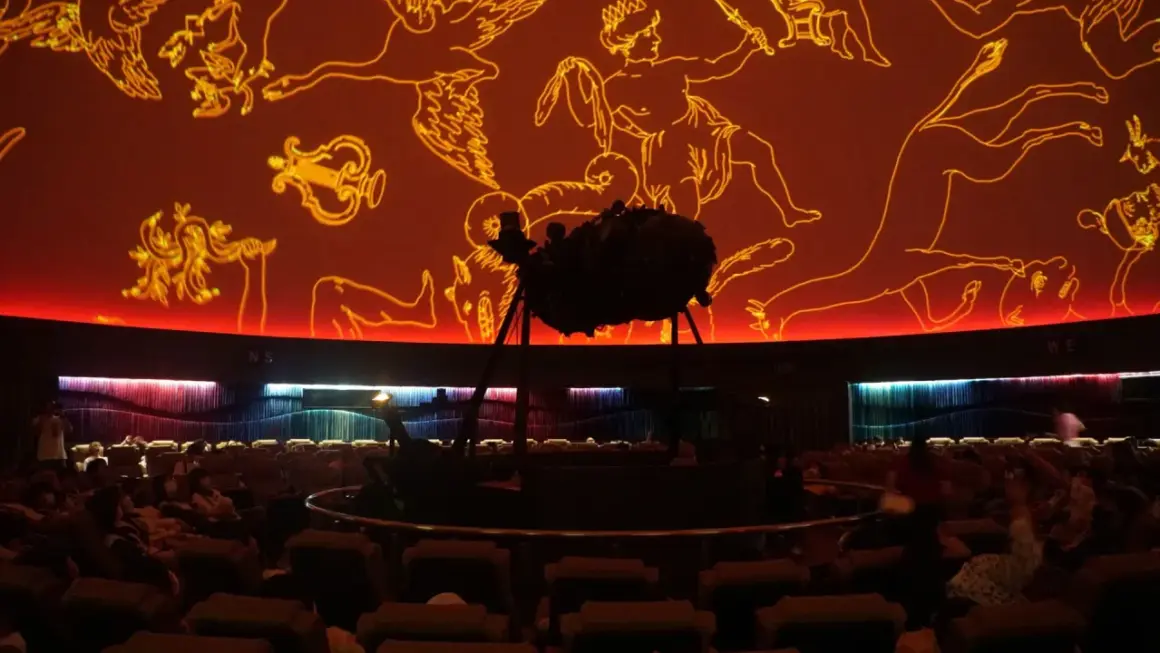 Wnętrze planetarium Warszawa. Na pomarańczowej kopule widnieją złote rysunki gwiazdozbiorów. Na środku stoi projektor.