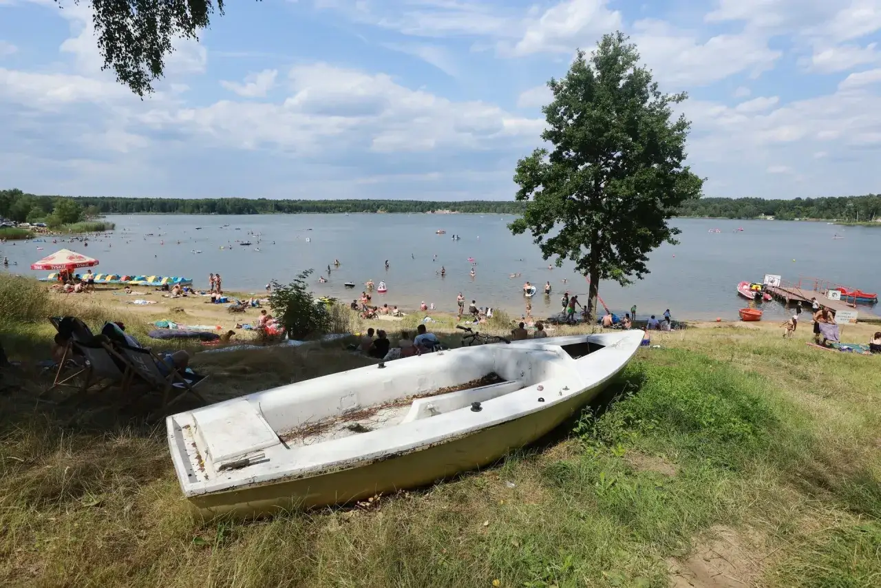 Letnia, słoneczna pogoda nad jeziorem. Ludzie odpoczywają na plaży i kąpią się w wodzie. Na pierwszym planie biała łódka, a w tle zielony las.