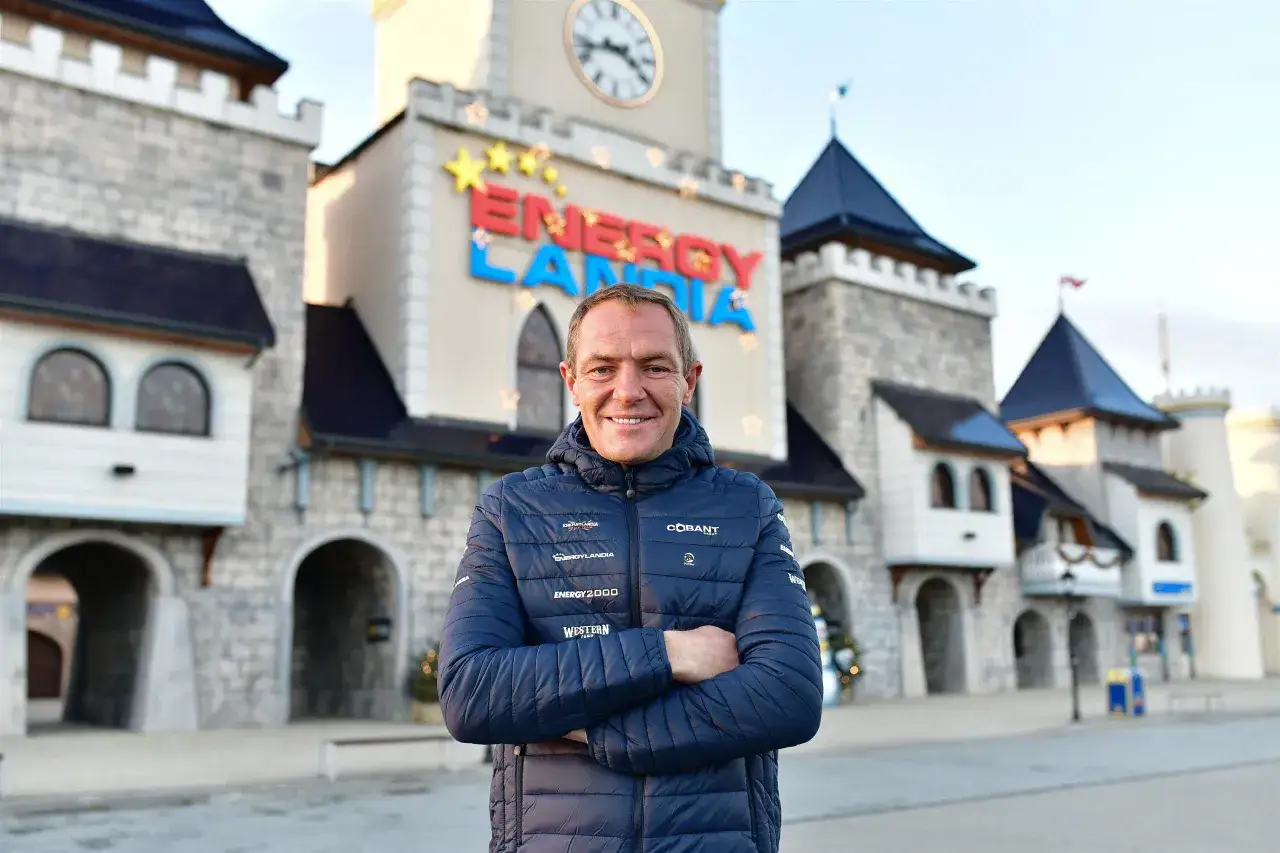 Właściciel Energylandii, uśmiechnięty mężczyzna w kurtce z logo parku, stoi przed bajkowymi budowlami.