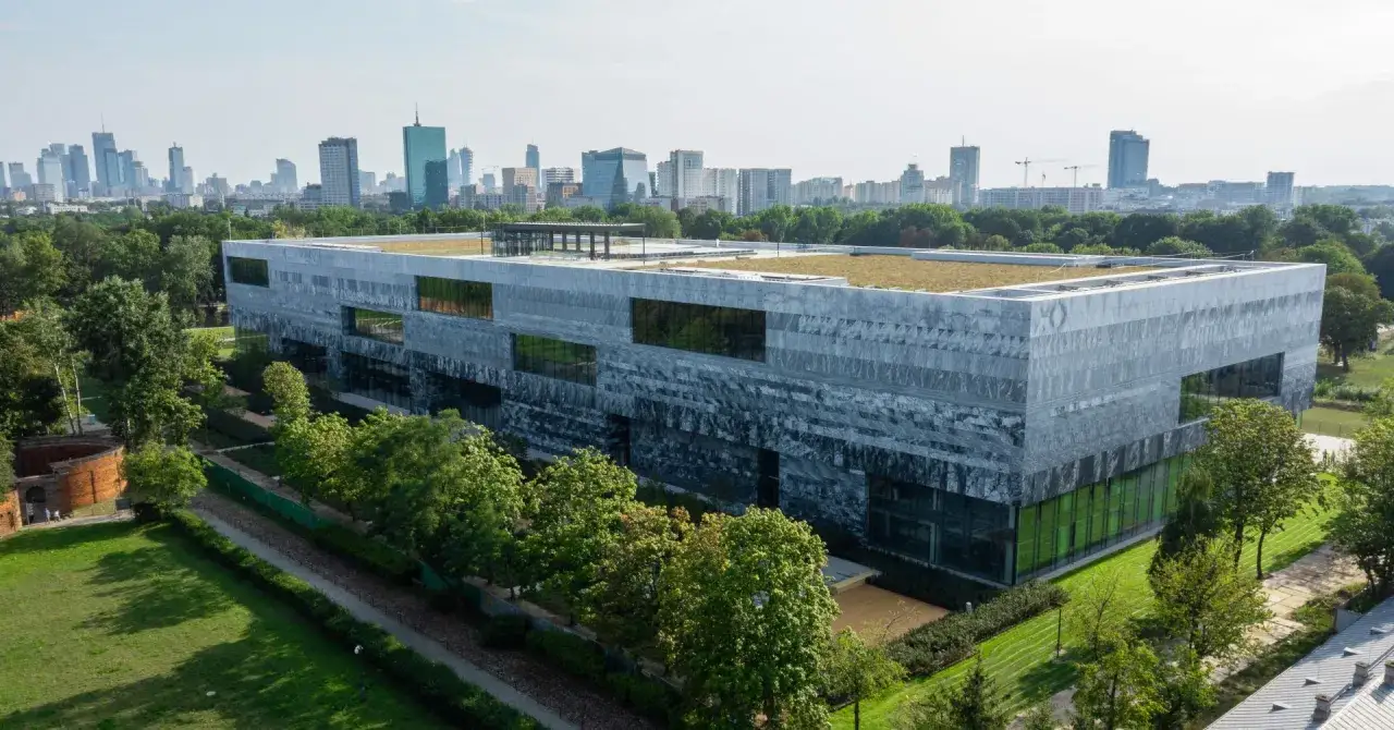 Nowoczesny budynek Muzeum Historii Polski w Warszawie, otoczony zielenią, z panoramą miasta w tle.