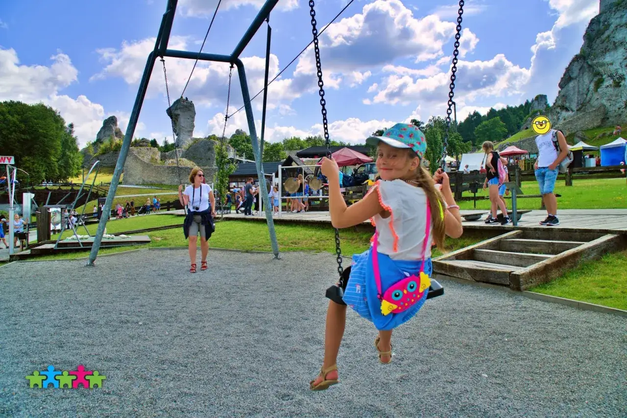 Dziewczynka na huśtawce w Ogrodzieńcu park rozrywki. W tle ruiny zamku i inni turyści.