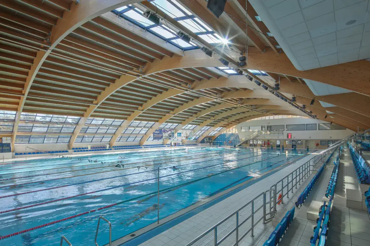 Basen sportowy w Warszawiance z drewnianą konstrukcją dachu i przeszklonymi oknami. Woda w basenie jest błękitna, a na torach pływają ludzie.