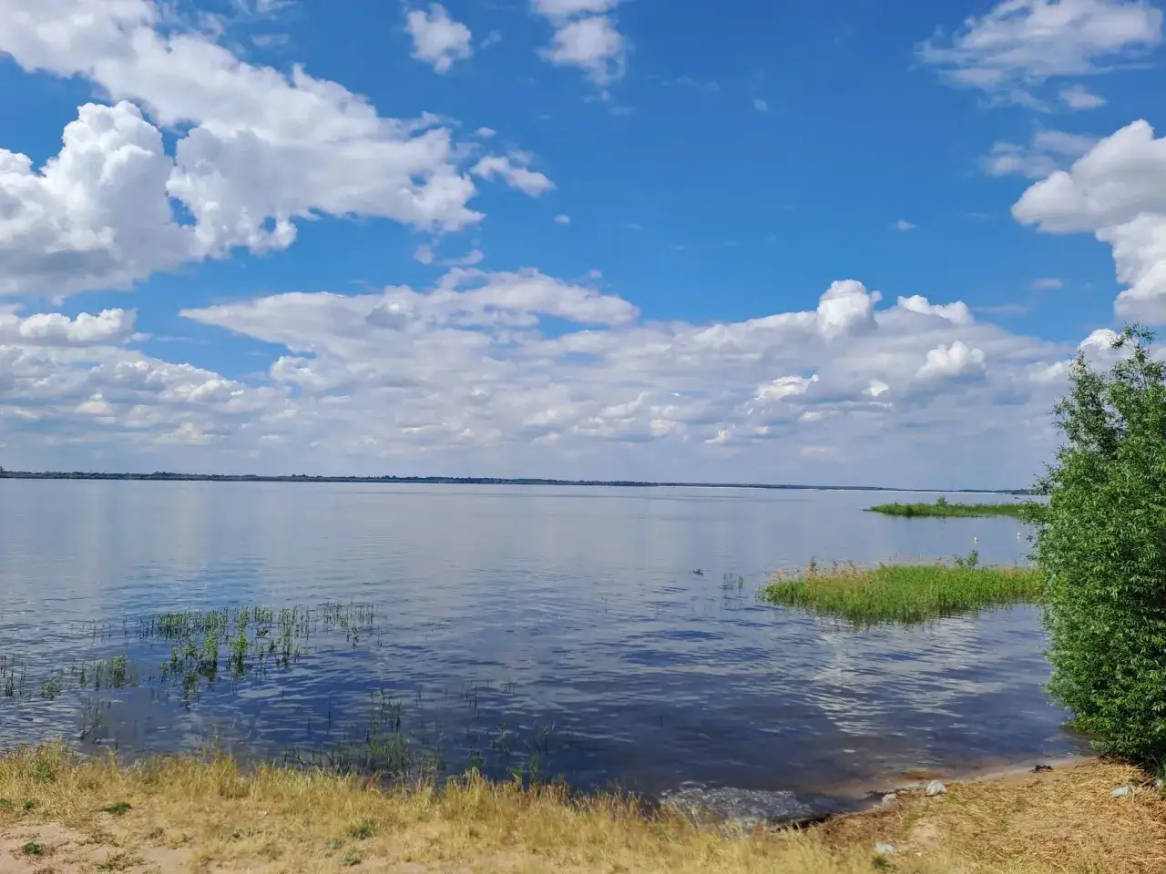 Spokojne jezioro pod błękitnym niebem z chmurami. Woda odbija niebo, a na brzegu rośnie trawa. Idealne miejsce na relaks i podziwianie jeziorsko atrakcje.