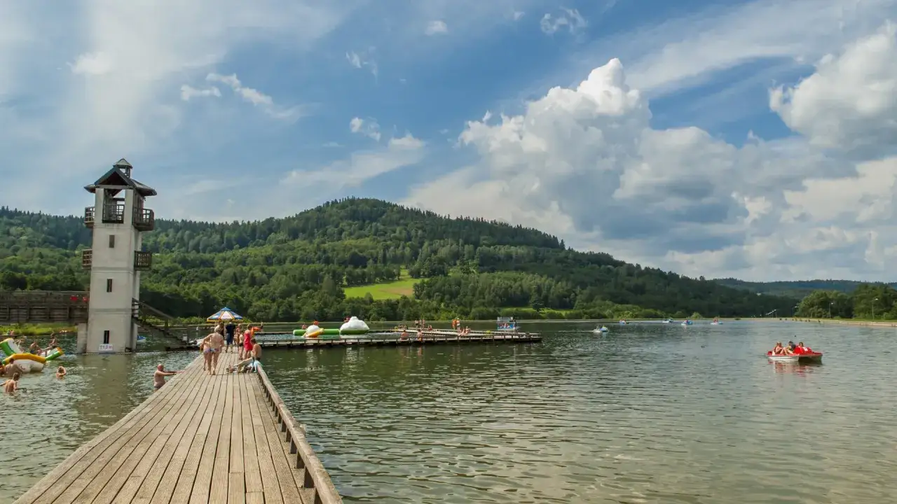 Ludzie wypoczywają nad wodą, na drewnianym molo i w łódkach. W tle widać wieżę i zalesione wzgórza. To idealne miejsce na letni relaks nad stara morawa zalew.