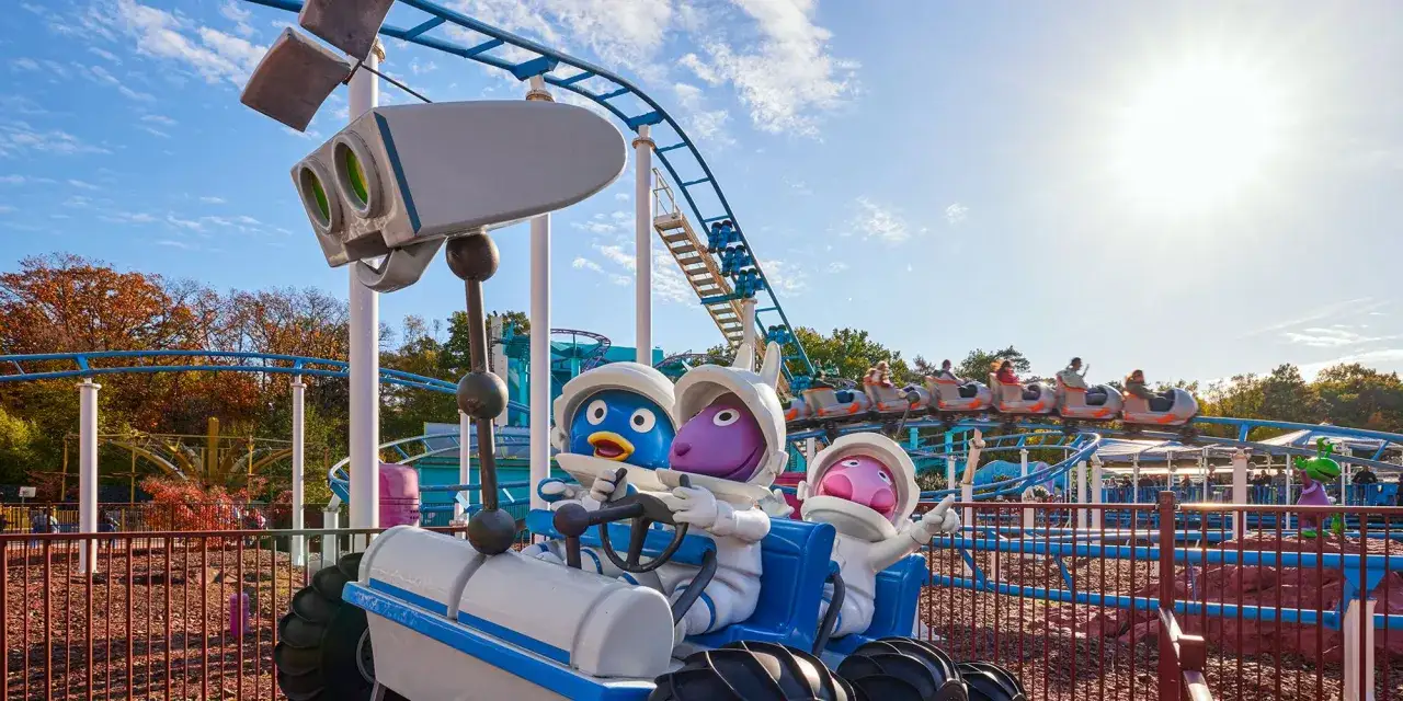 Trzy postacie z kreskówek w skafandrach kosmicznych jadą traktorem na tle kolejki górskiej w Movie Park Germany.