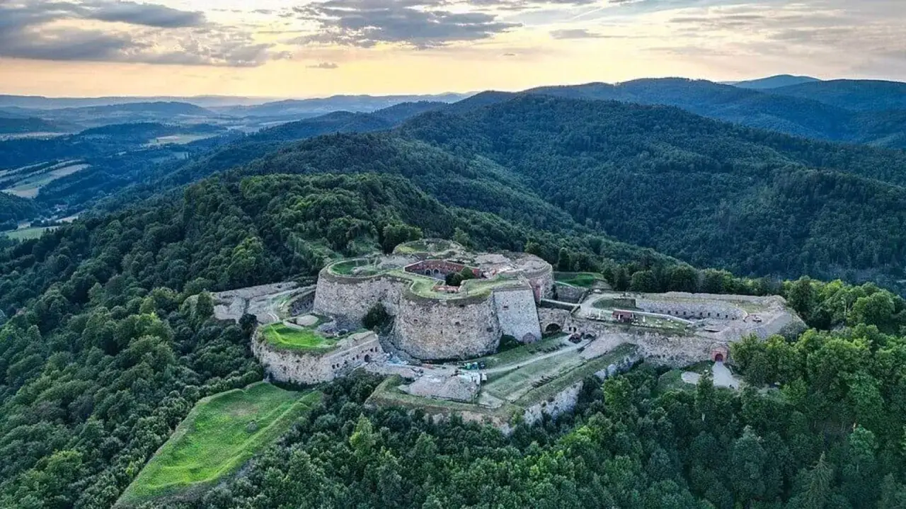 Twierdza na zielonej górze, otoczona lasami, niczym srebrna korona na tle zachodzącego słońca.