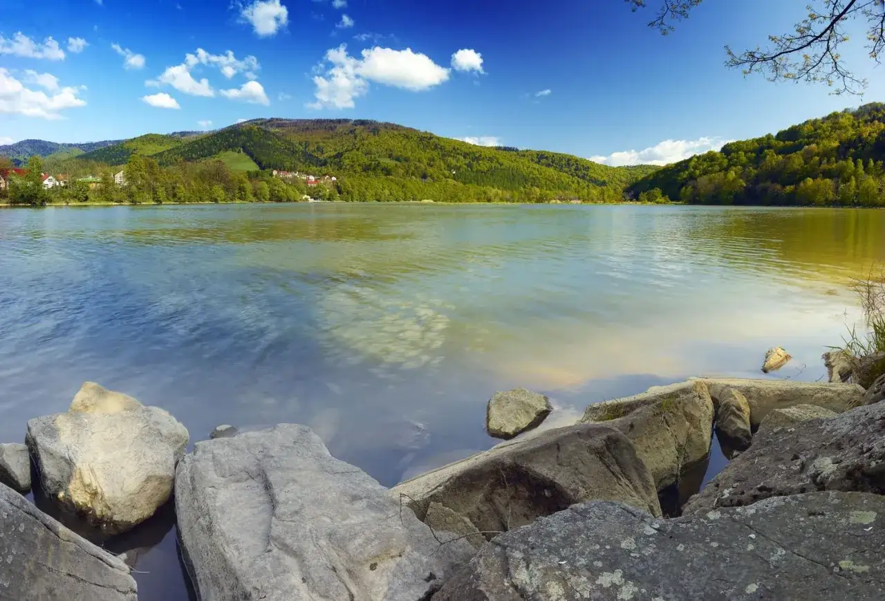 Kamienista plaża nad Jeziorem Międzybrodzkim, otoczona zielonymi wzgórzami pod błękitnym niebem z chmurami.