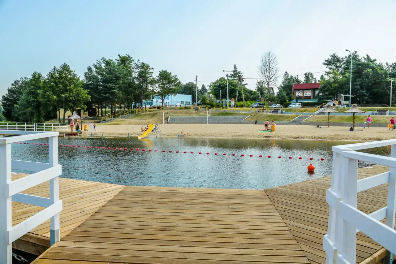 Słoneczny dzień nad zalewem Siemiatycze. Ludzie odpoczywają na piaszczystej plaży, dzieci bawią się na zjeżdżalni.