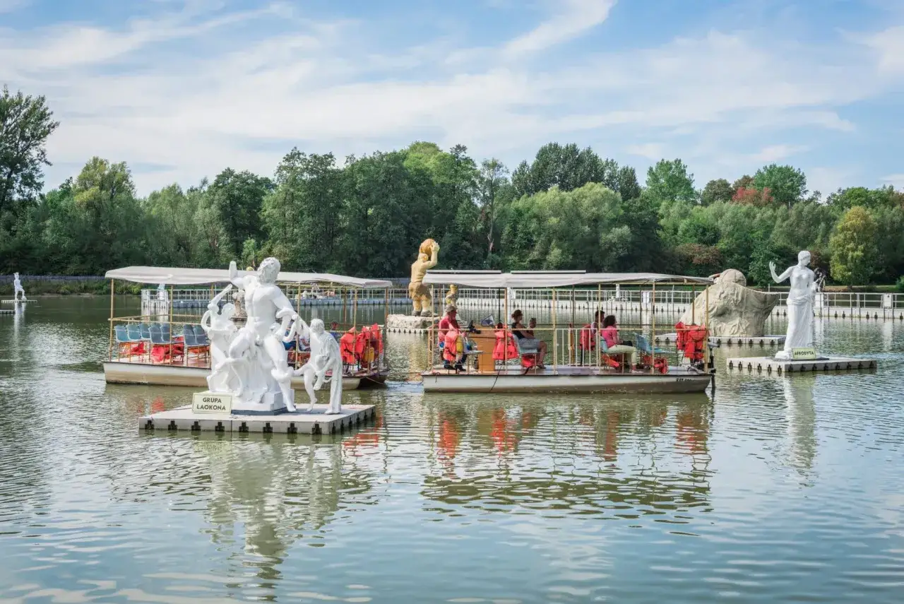 Pływające platformy z rzeźbami mitologicznymi tworzą niezwykły park mitologii na wodzie. Ludzie podziwiają dzieła sztuki z łodzi.