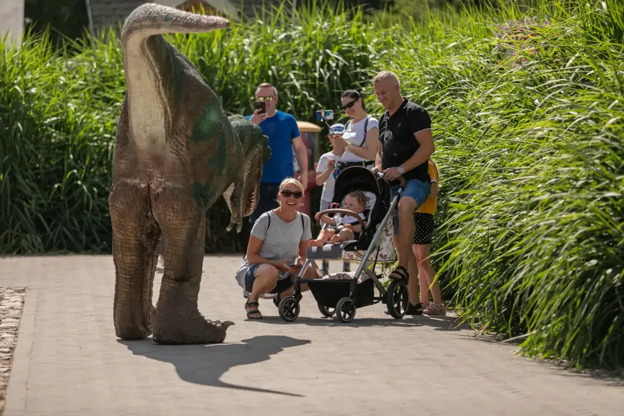 Rodzina podziwia dinozaura w parku ewolucji w Sławutówku. Dziecko w wózku z zaciekawieniem obserwuje prehistoryczne stworzenie.