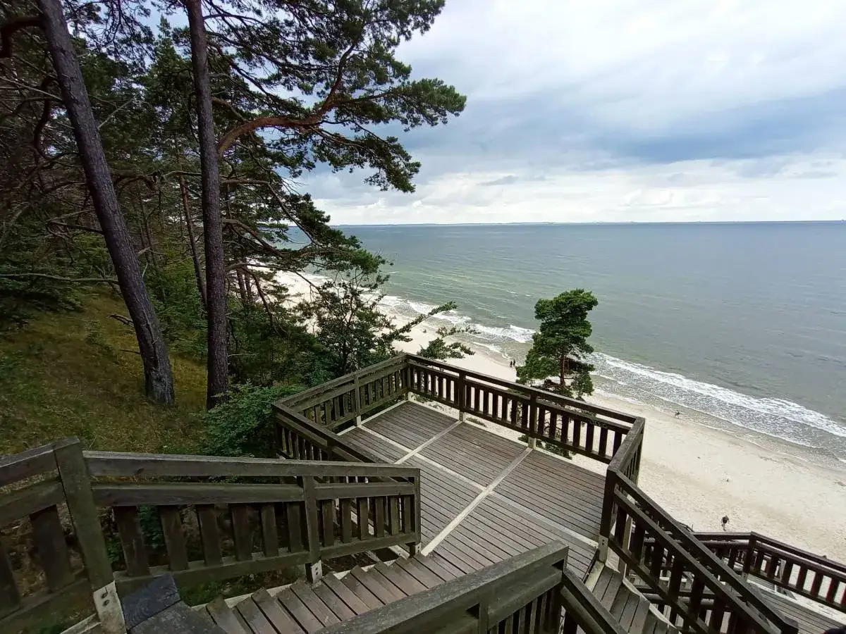 Drewniane schody prowadzą na plażę z widokiem na morze. W oddali widać las, a całość przypomina krajobraz z Kawczej Góry.