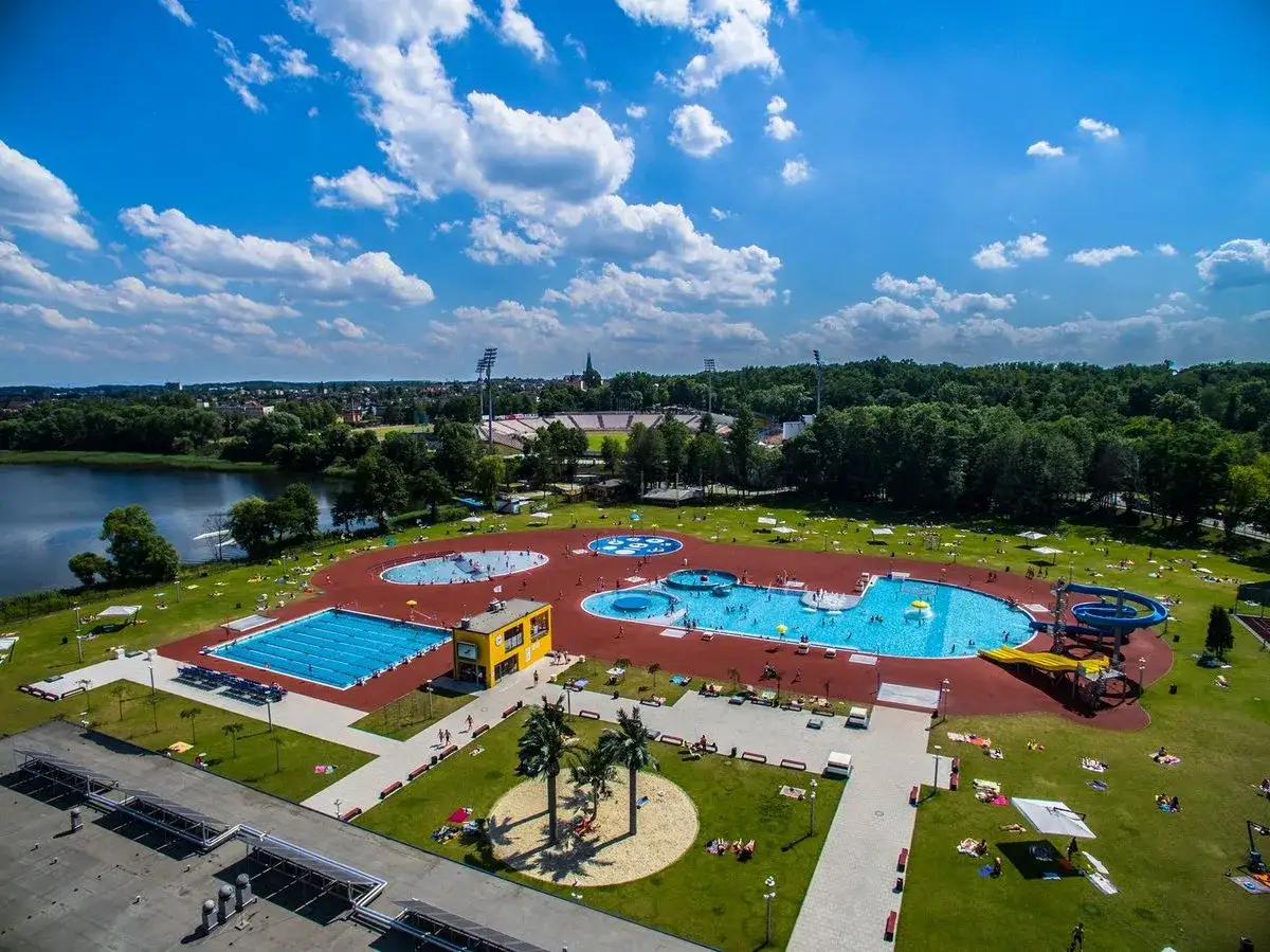 Letni dzień nad wodą. Widać rozległy kompleks basenów z wodnymi atrakcjami, zjeżdżalniami i leżakami. W tle jezioro i stadion.