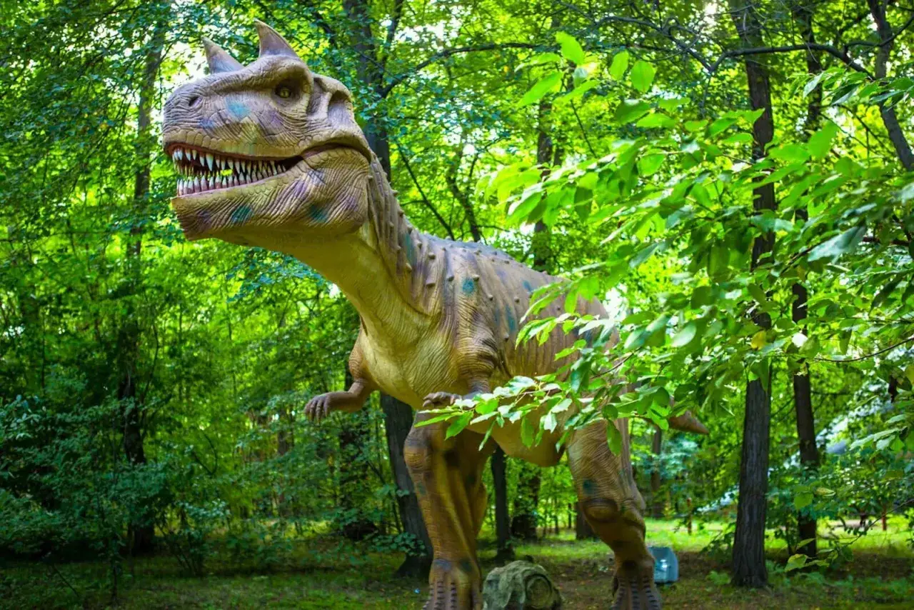 Witaj w parku ruchomych dinozaurów! Gigantyczny, realistyczny model tyranozaura z otwartą paszczą i ostrymi zębami czeka na Ciebie wśród bujnej zieleni.