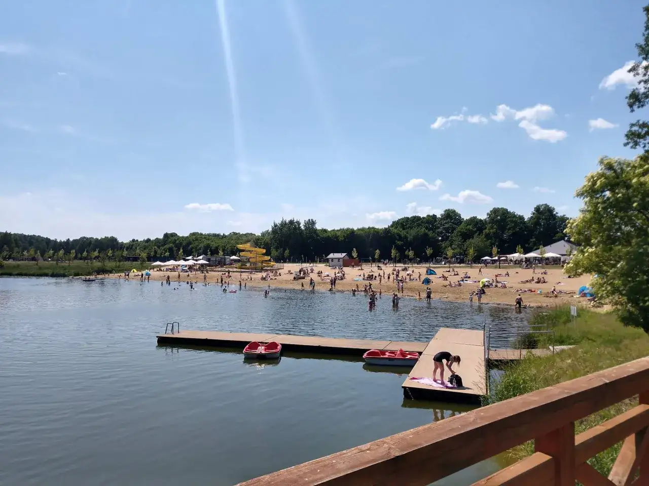 Słoneczny dzień nad zalewem Lisowice. Ludzie odpoczywają na plaży, kąpią się i pływają łódkami.