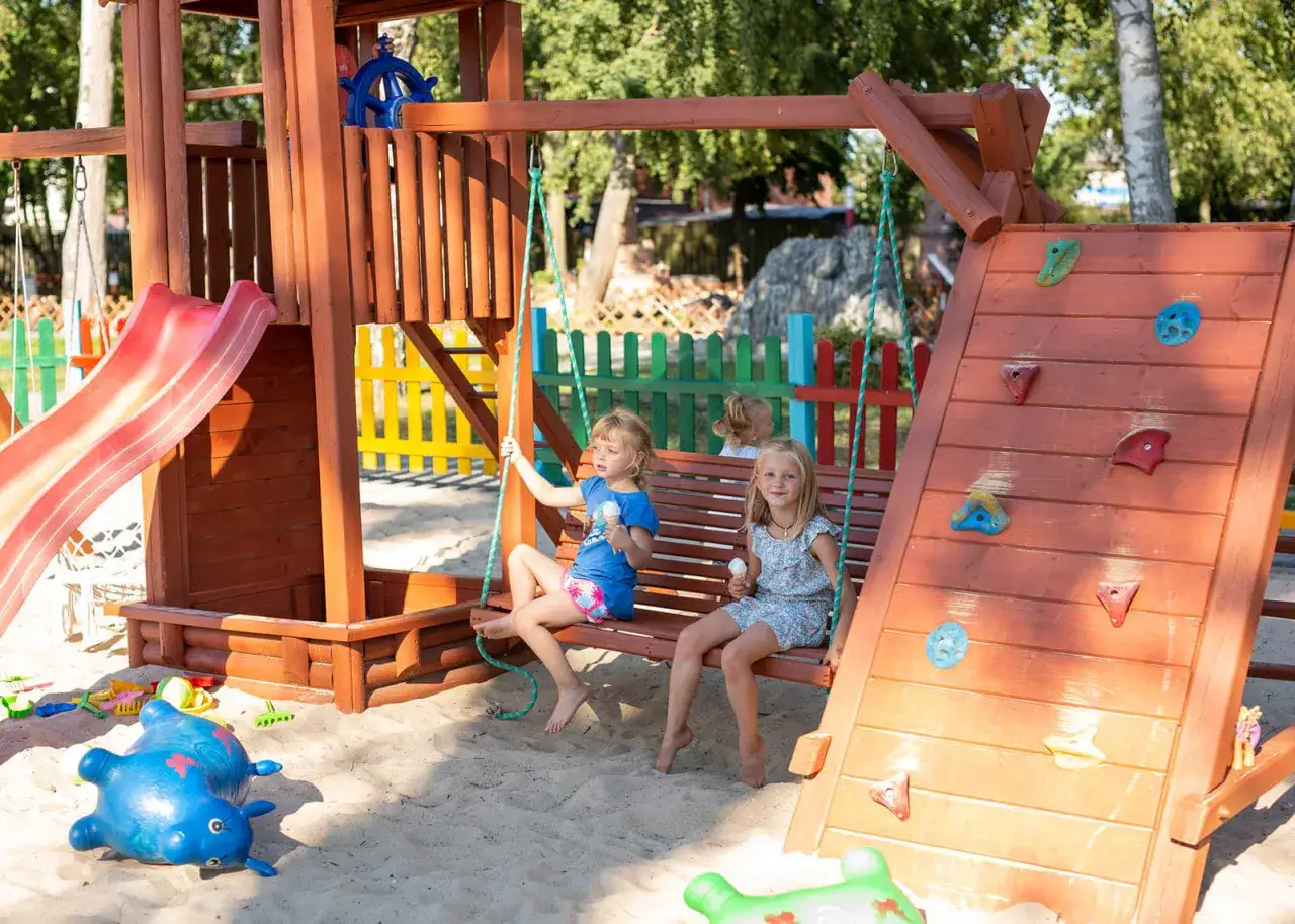 Dwie dziewczynki jedzą lody na huśtawce w piaskownicy. W tle zjeżdżalnia i ścianka wspinaczkowa. To idealne miejsce na zabawę w parku miniatur i kolejek dziwnów.