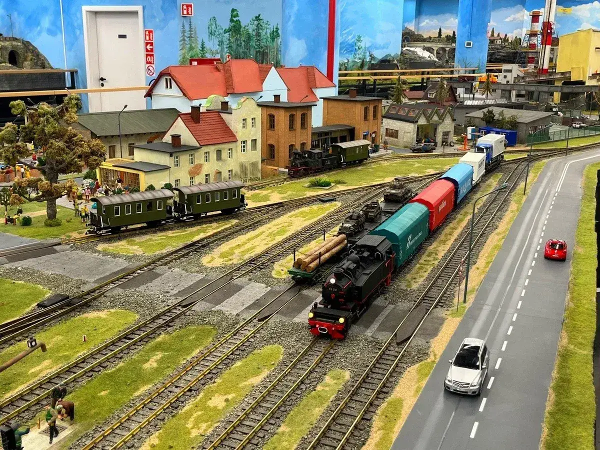 Miniaturowa kolejkowo w Warszawie z parowozem i wagonami. W tle miasteczko z domkami i fabryką.