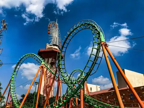 Zielona kolejka górska z pomarańczowymi podporami, w tle wieża widokowa i diabelski młyn. Ten tilt coaster zapowiada emocjonującą przejażdżkę.