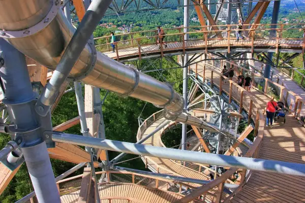 Drewniana konstrukcja z metalową zjeżdżalnią tworzy ścieżkę w koronach drzew. Ludzie spacerują po platformach, podziwiając widoki. To wspaniały sky walk!