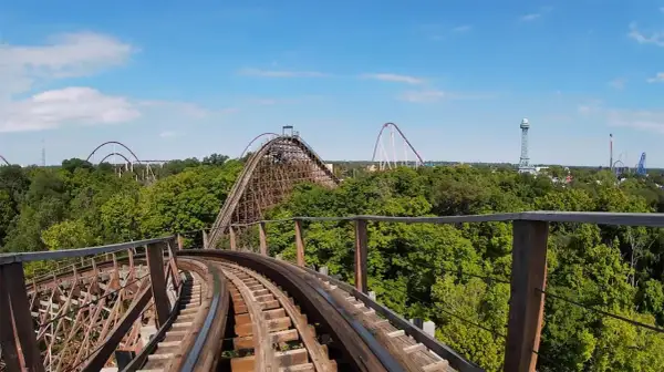 Widok z kolejki górskiej na tle zielonych drzew i błękitnego nieba. W oddali widać inne atrakcje parku rozrywki, w tym największy drewniany rollercoaster na świecie.