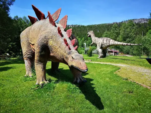 W parku dinozaurów karłów Stegozaur pochyla się do trawy, podczas gdy Tyranozaur czuwa w oddali.
