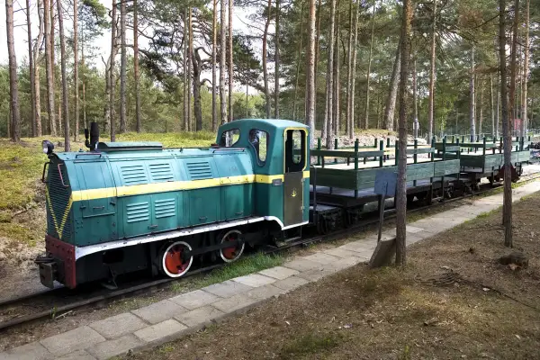 Zielona lokomotywa z żółtym pasem i wagonami na torach leśnych. Ekspozycja w Muzeum Obrony Wybrzeża w Helu.