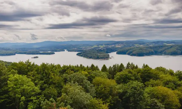 Widok na jezioro solińskie otoczone zielonymi wzgórzami i lasami pod pochmurnym niebem.