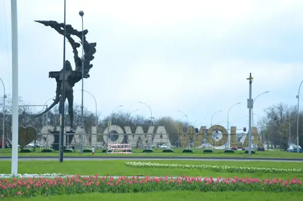 Pomnik i napis "STALOWA WOLA" w parku, otoczone kwitnącymi tulipanami.