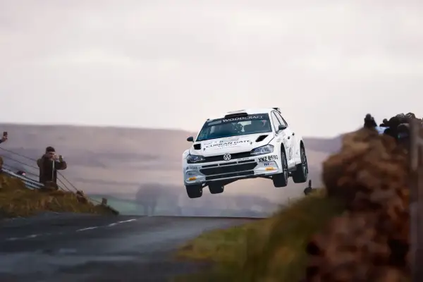 Biały samochód rajdowy zespołu Energylandia Rally Team w locie nad wzniesieniem. Widzowie filmują akcję.