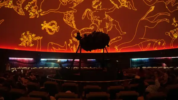 Wnętrze planetarium Warszawa. Na pomarańczowej kopule widnieją złote rysunki gwiazdozbiorów. Na środku stoi projektor.