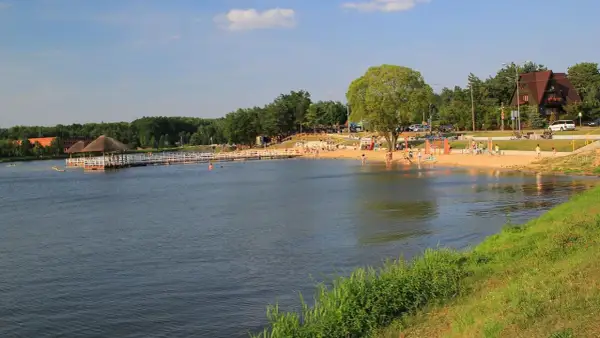 Słoneczny dzień nad jeziorem w Siemiatyczach. Plaża pełna ludzi, pomost z altaną, a w tle las i budynki. Idealne siemiatycze atrakcje.