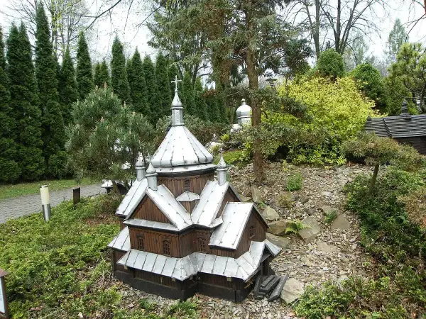 Drewniana makieta cerkwi z metalowym dachem, część parku miniatur centrum kultury ekumenicznej, otoczona zielenią i kamieniami.