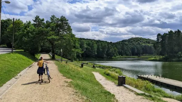 Kobieta z rowerem na ścieżce nad jeziorem, otoczonym lasem. W oddali widać wzgórza, a na wodzie pływają kaczki. To idealne miejsce na spacer, jak złoty potok.