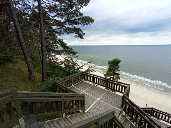 Drewniane schody prowadzą na plażę z widokiem na morze. W oddali widać las i **kaczą górę**.