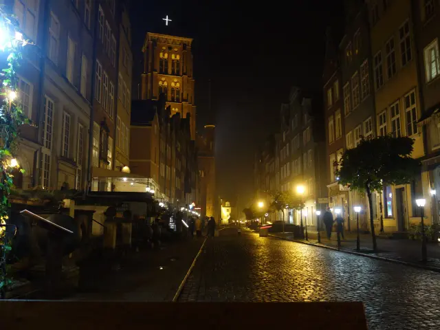 Mroczny, brukowany zaułek w Gdańsku nocą. Latarnie rzucają złote światło na mokrą nawierzchnię, a w oddali widać wieżę kościoła.