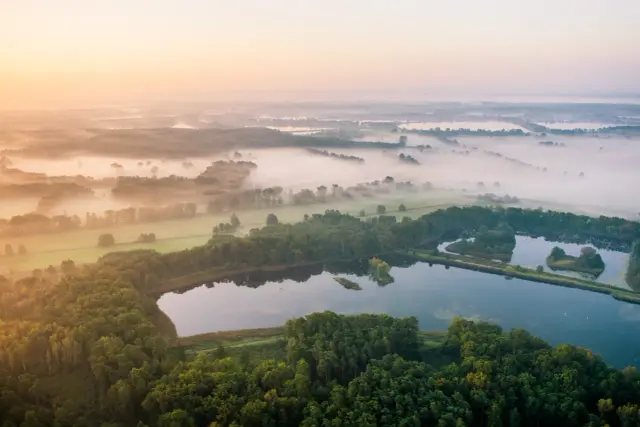 Mglisty poranek nad stawami w Dolinie Baryczy. Widok z lotu ptaka na lasy, pola i wodę, ukazujący piękno przyrody i liczne atrakcje tego regionu.